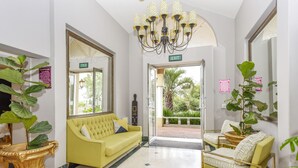 A lobby with a yellow sofa, a green sofa, and a yellow armchair. There is a chandelier, a mirror, and a plant.