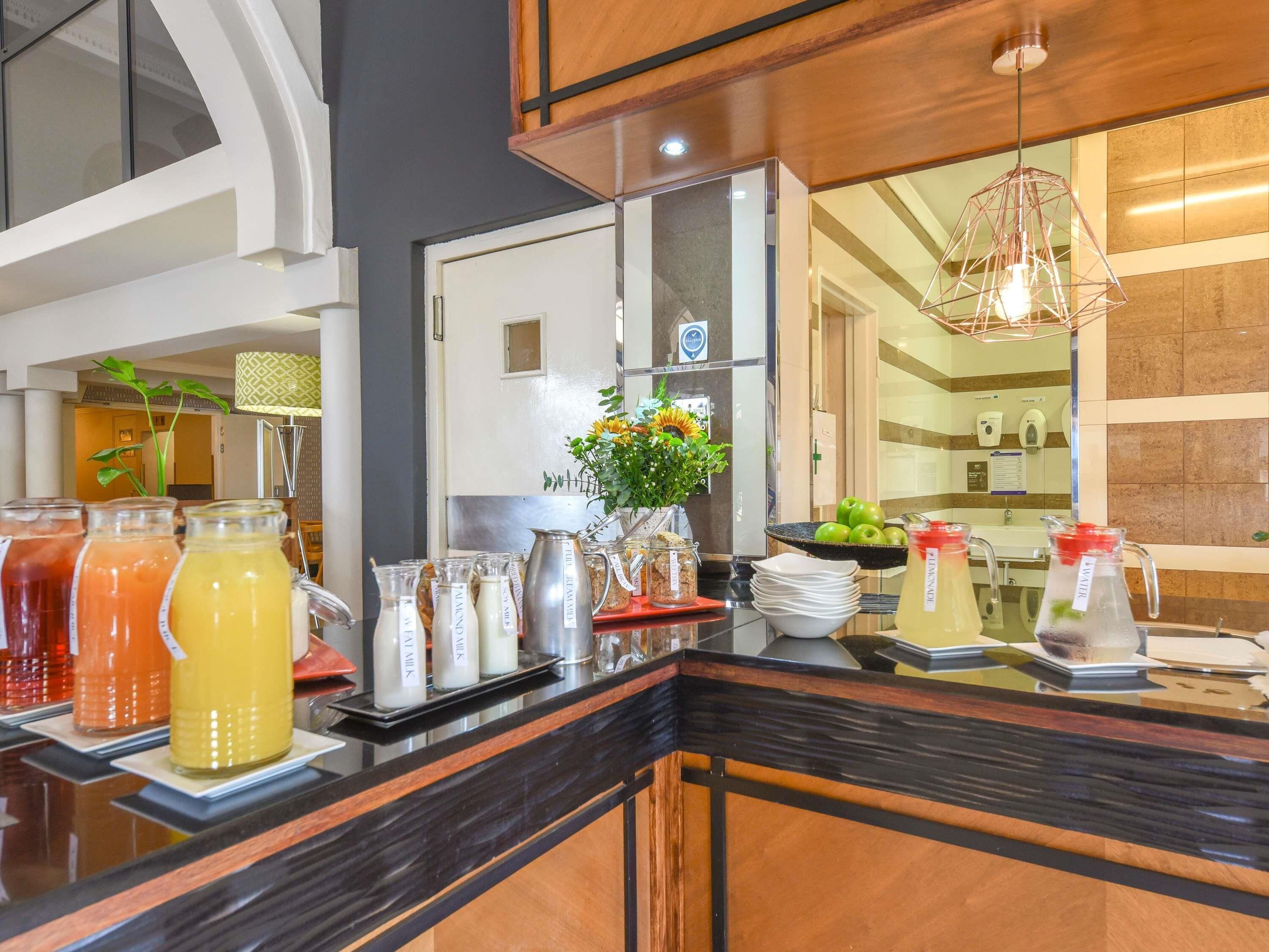Zona de recepción de un hotel con un mostrador que exhibe diversas bebidas y alimentos, un espejo y una planta decorativa.