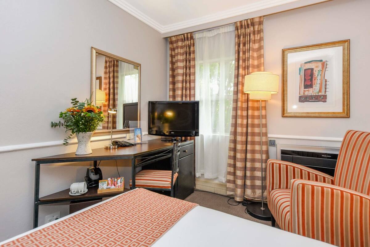 Een hotelkamer met een bed, een bureau met een televisie, een lamp, een stoel en een schilderij aan de muur.