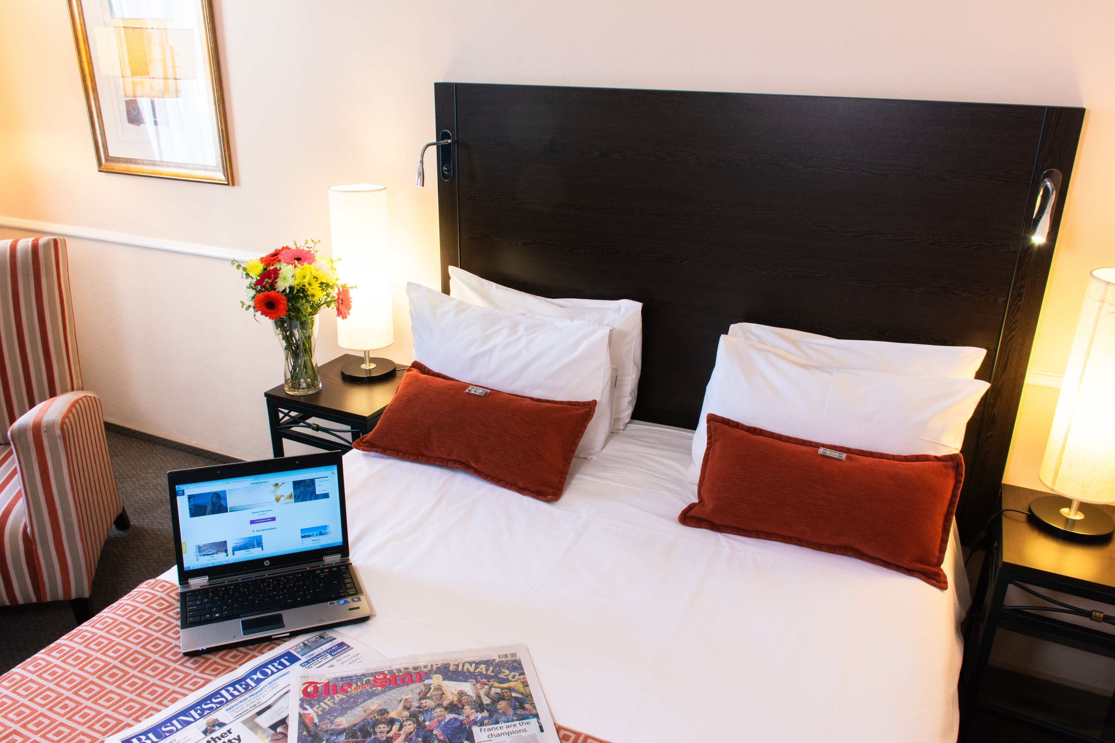 A hotel room with a bed, a laptop, a magazine, and a vase with flowers.