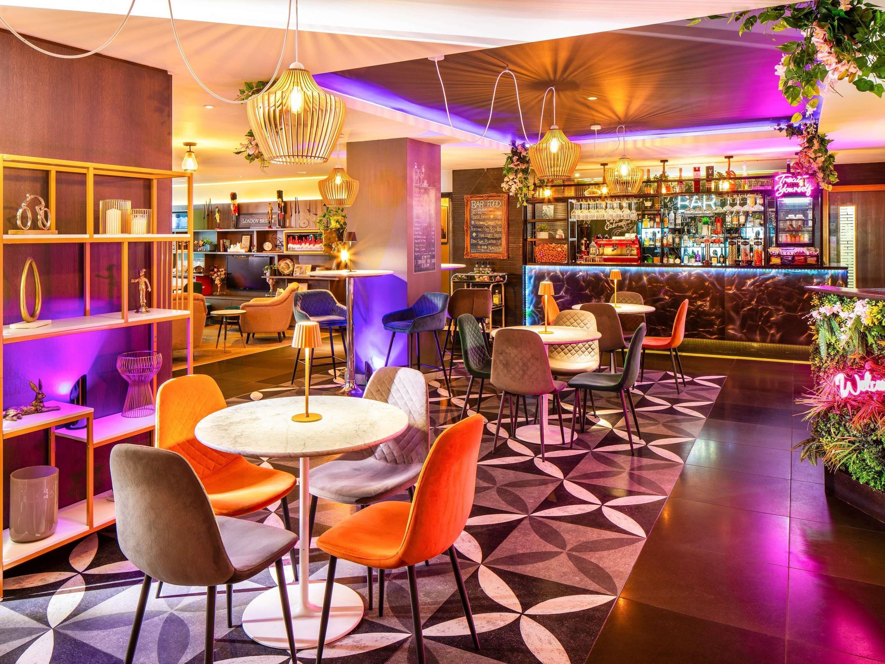 Uma área de bar moderna com mesas redondas e cadeiras laranja, um balcão com letreiro de neon e prateleiras com diversos objetos.