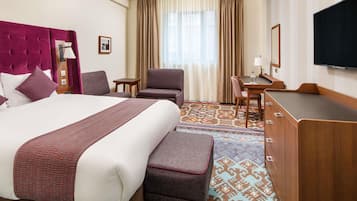 Habitación de hotel con una cama grande, un escritorio, una silla, un televisor y una alfombra estampada.
