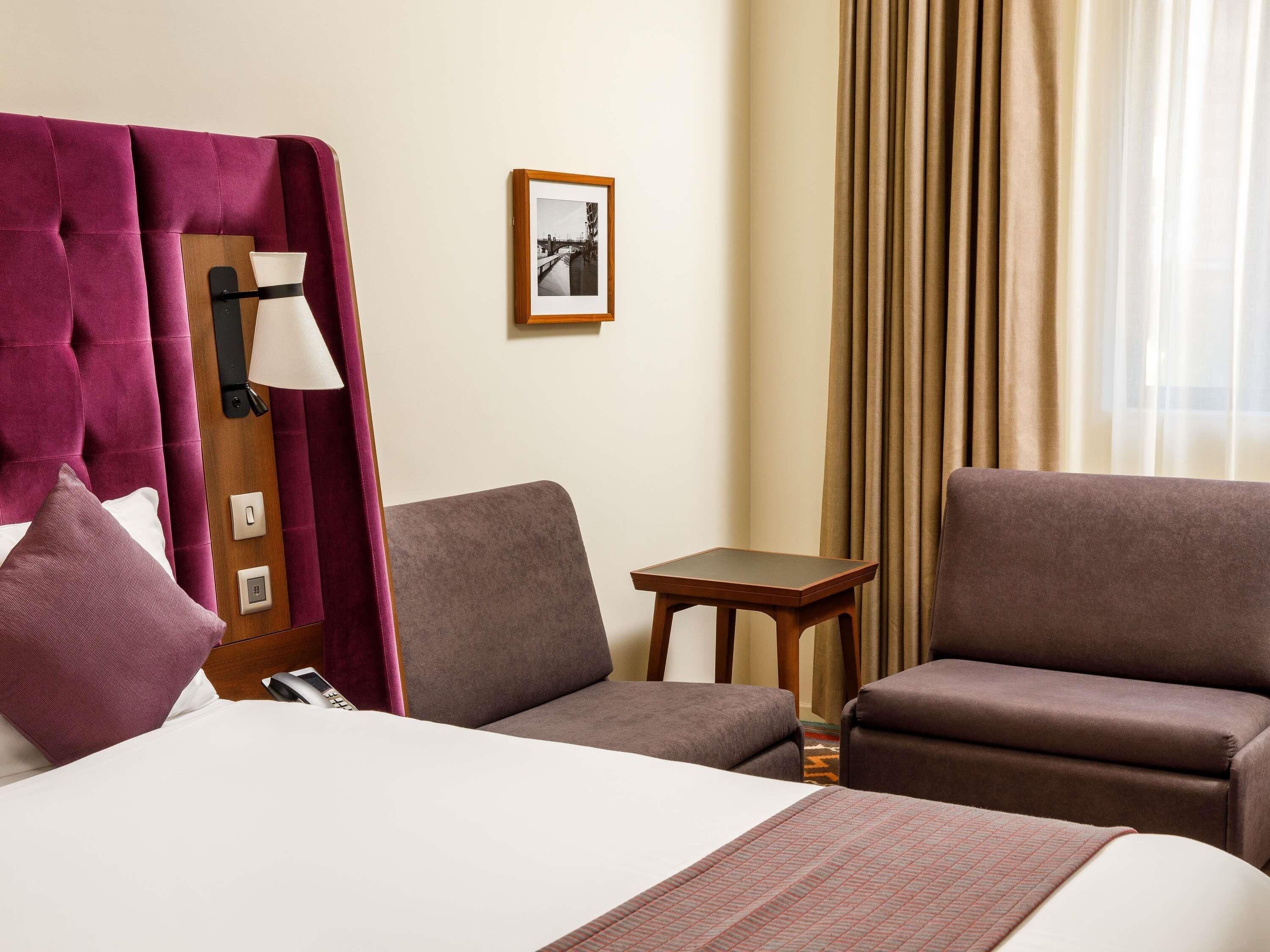 A hotel room with a bed, two armchairs, a small table, a lamp, a picture on the wall, and curtains.