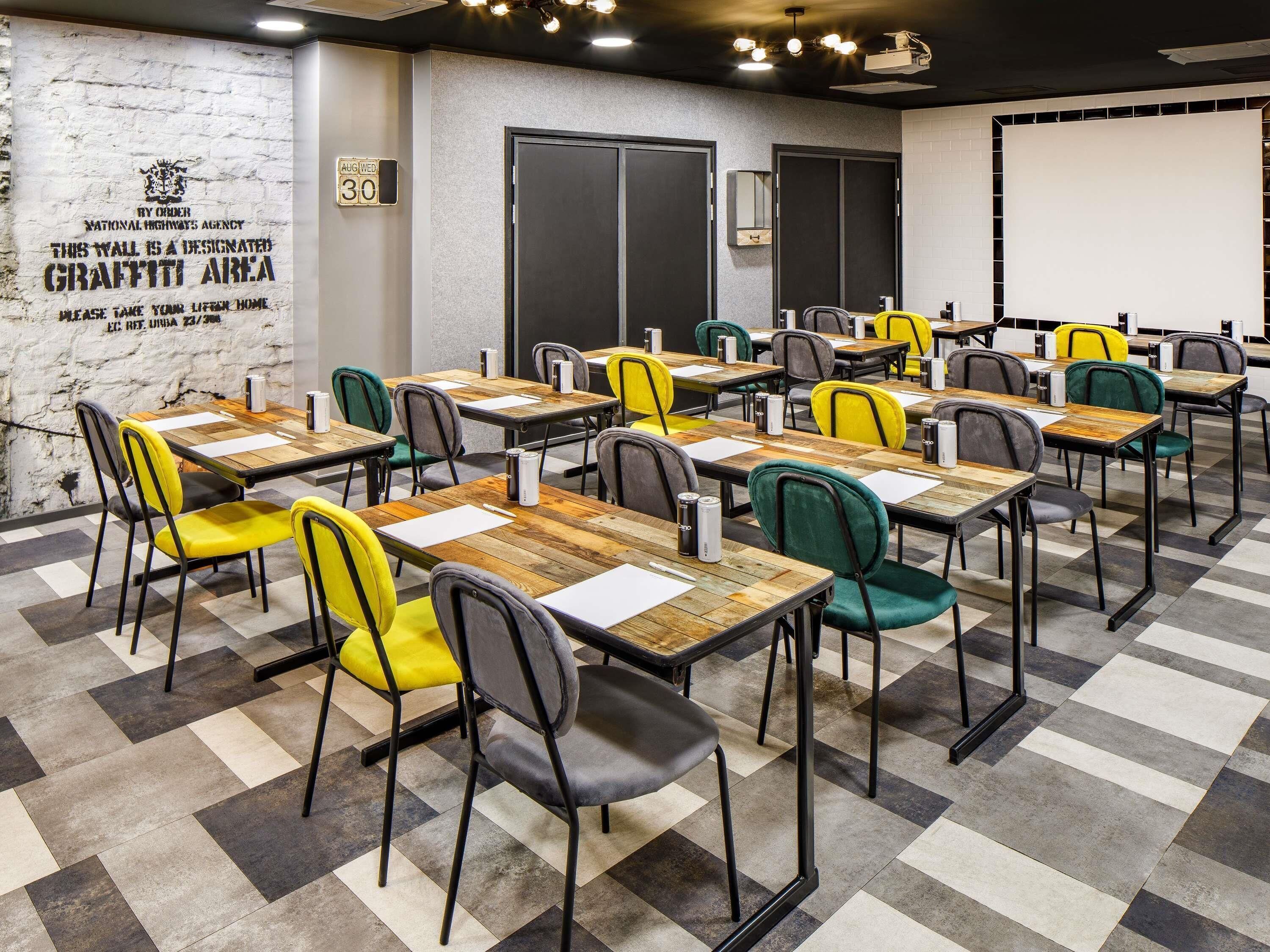 A conference room with tables and chairs, a projector screen, and a wall with a graffiti area sign.