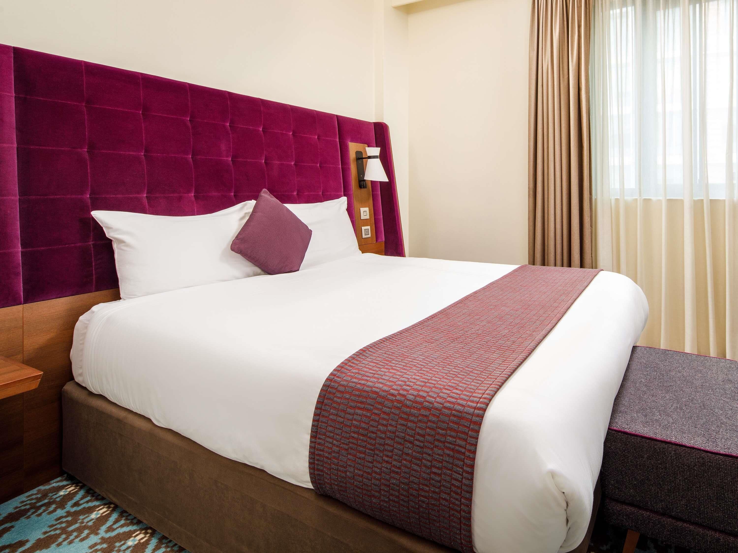 A hotel room with a large bed, a headboard, a nightstand, and a window with curtains.