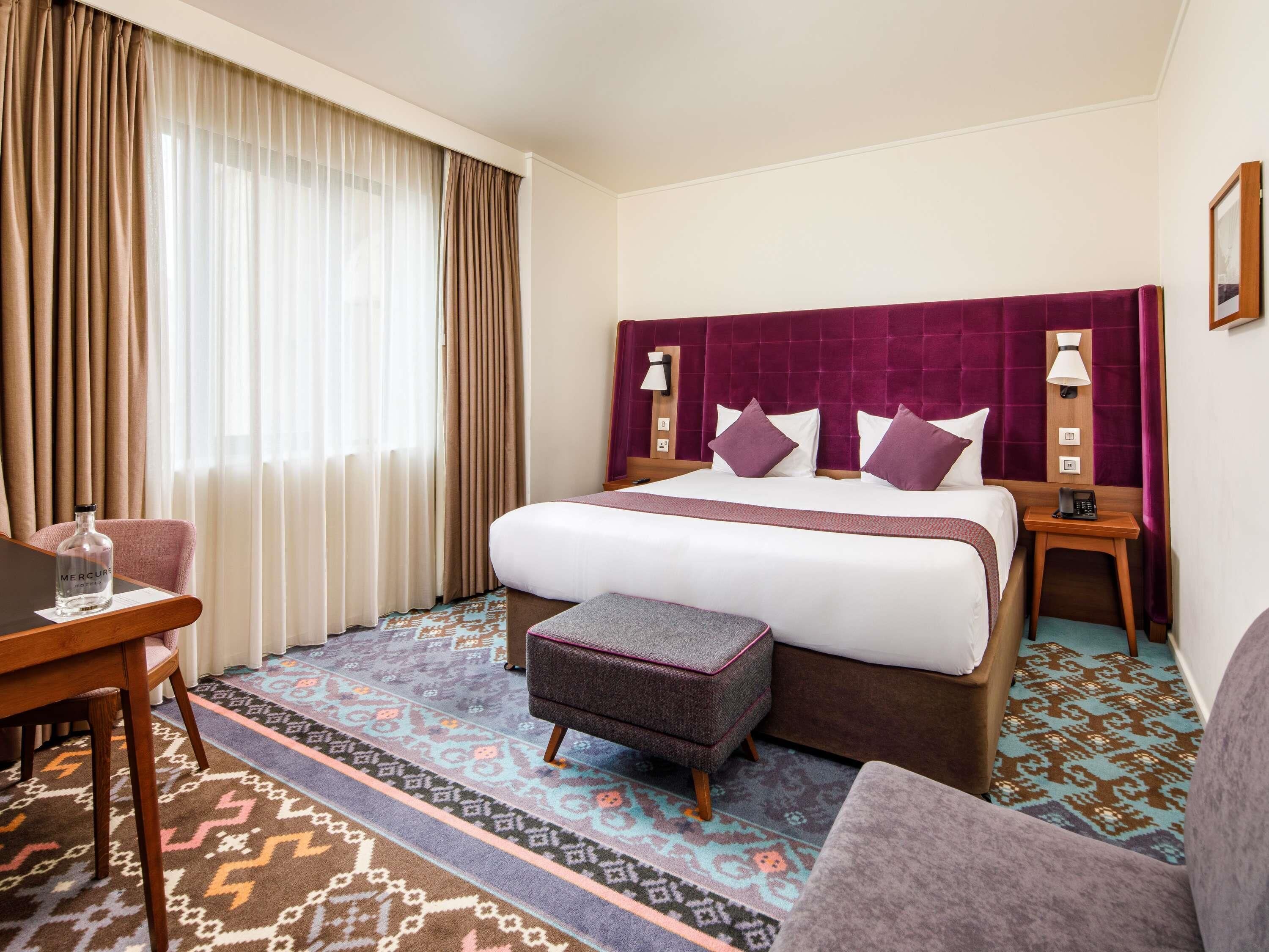 A hotel room with a bed, a desk, a chair, a small table, and a window with curtains.