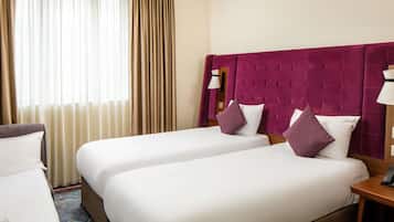 Ein Hotelzimmer mit zwei Betten, einem großen Fenster mit Vorhängen, einem violetten Kopfteil und einem Nachttisch mit Lampe.