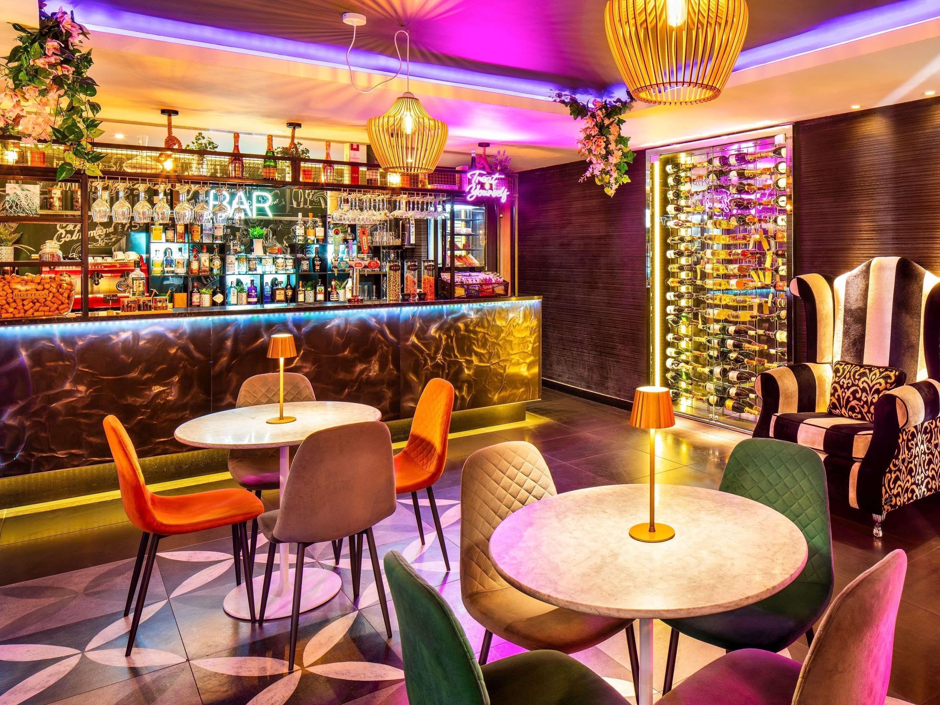 Uma área de bar com variedade de garrafas e copos, assentos com mesas redondas, e uma parede com garrafas de vinho.