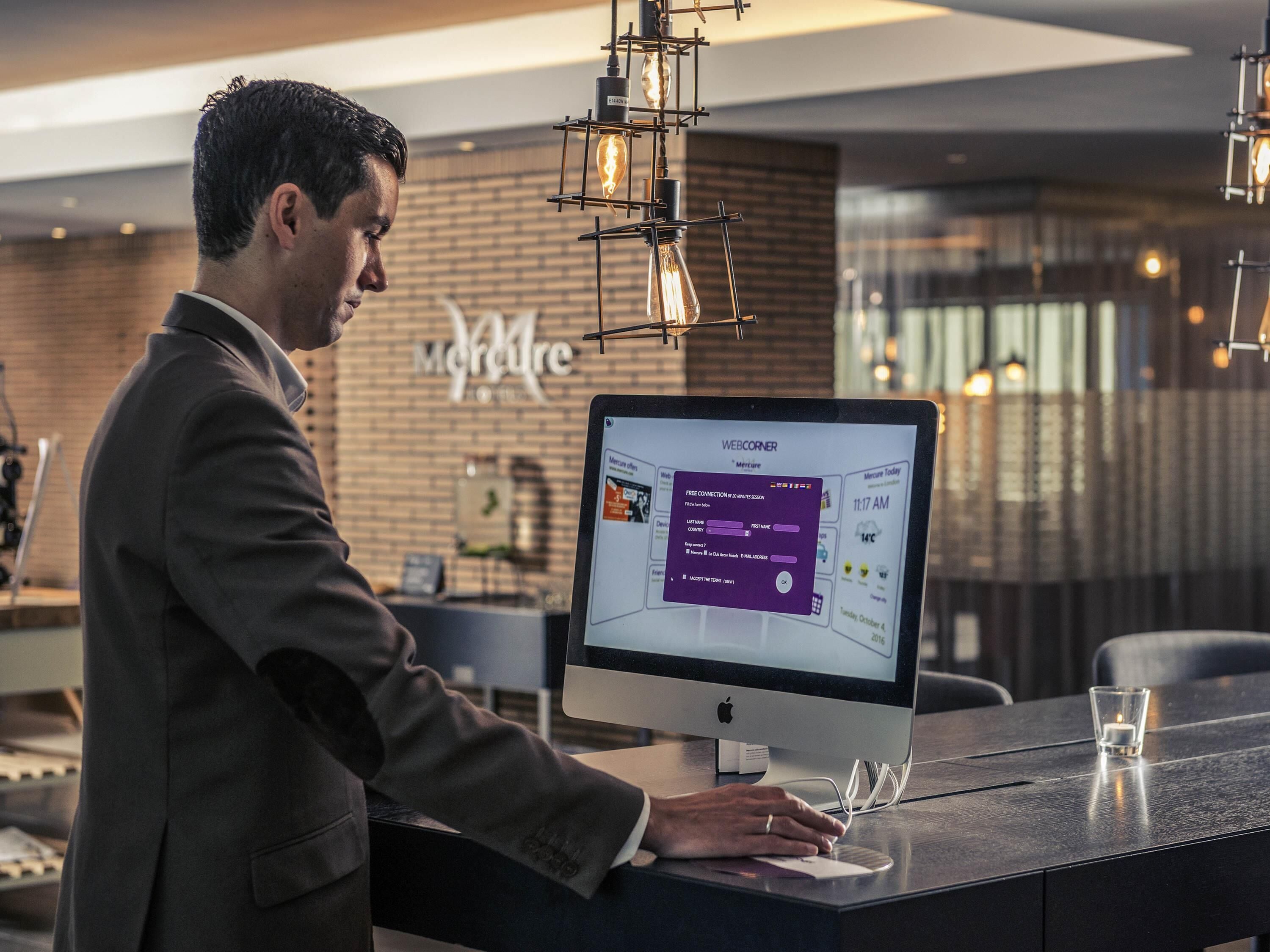 A man at a desk with a computer displaying a booking interface, in a modern hotel lobby with a brick wall and pendant lighting.