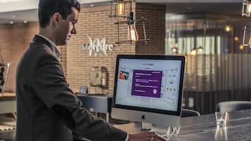 A man at a desk with a computer displaying a booking interface, in a modern hotel lobby with a brick wall and pendant lighting.