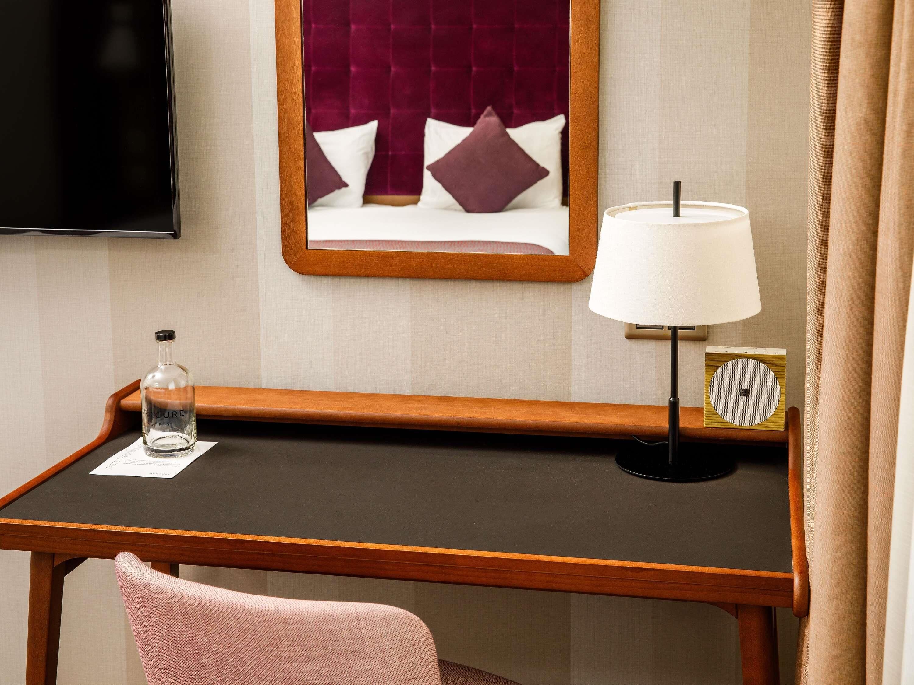 Quarto de hotel com uma escrivaninha de madeira, televisão na parede, cama com cabeceira vermelha, luminária de cabeceira, um pequeno alto-falante e uma garrafa de água na escrivaninha.