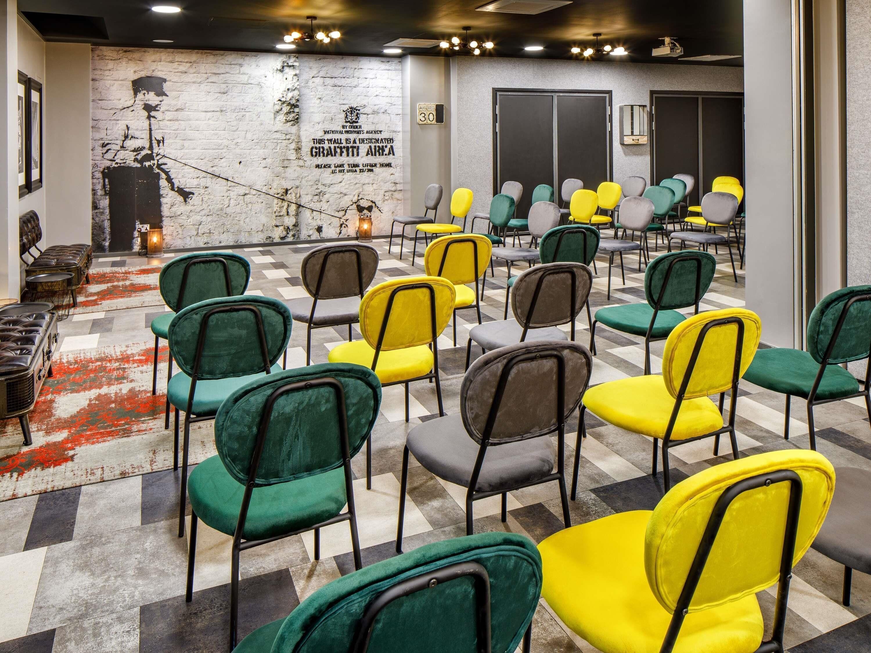 Uma sala de conferências com piso xadrez, cadeiras verdes e amarelas, um muro com grafites e uma lareira.