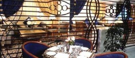 Un espace repas agrémenté d’une cloison en métal à motifs, de chaises recouvertes d’un tissu bleu et d’une petite table ronde recouverte d’une nappe blanche et ornée d’un vase de fleurs.