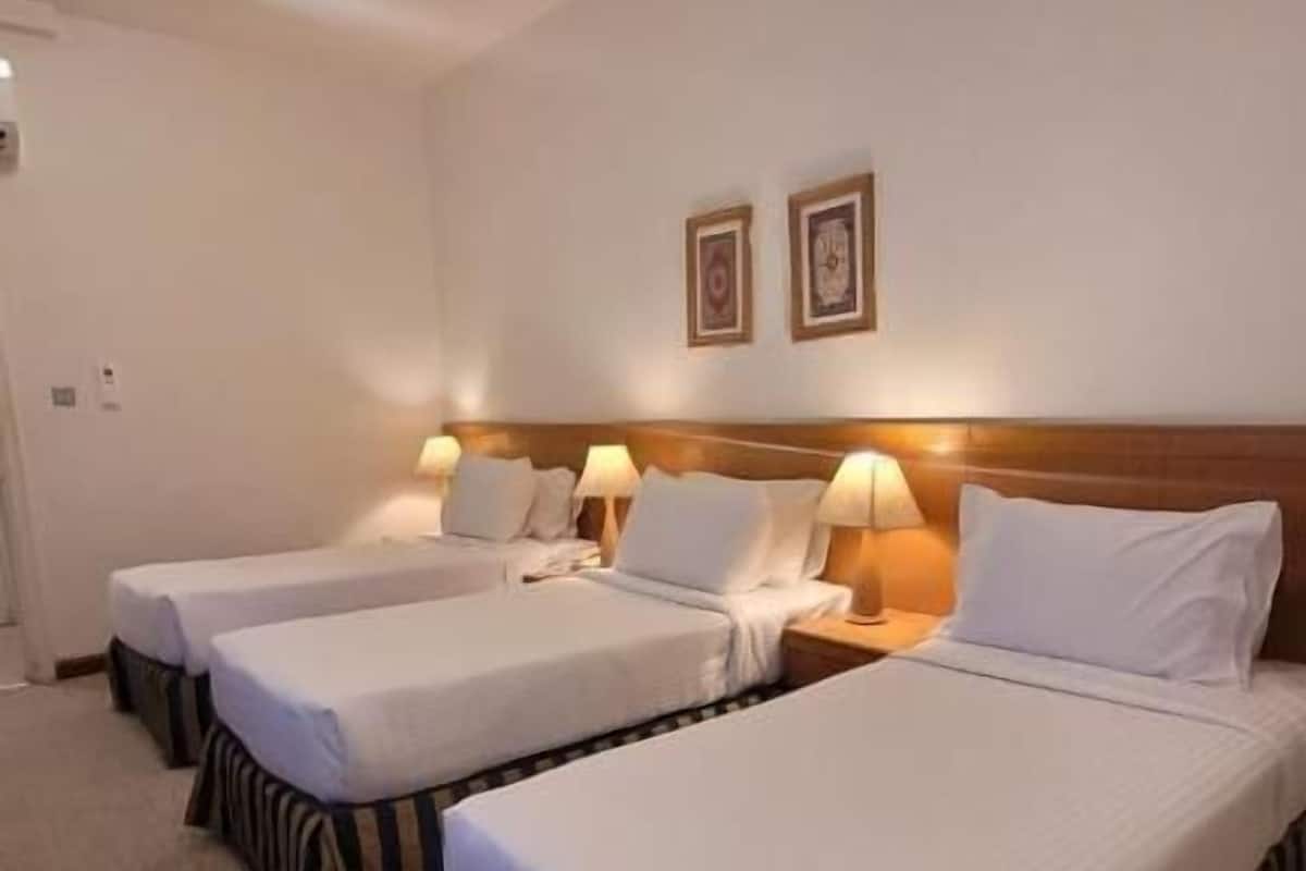 A hotel room with three single beds, each with a wooden headboard and white bedding. There are two bedside lamps and two framed pictures on the wall.