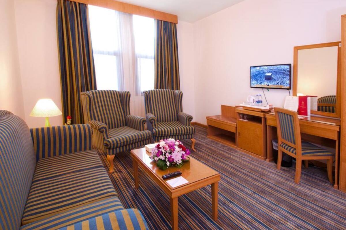 A hotel room with a striped sofa, two striped armchairs, a wooden coffee table with a vase of flowers, a desk with a chair, a television, and a large mirror.