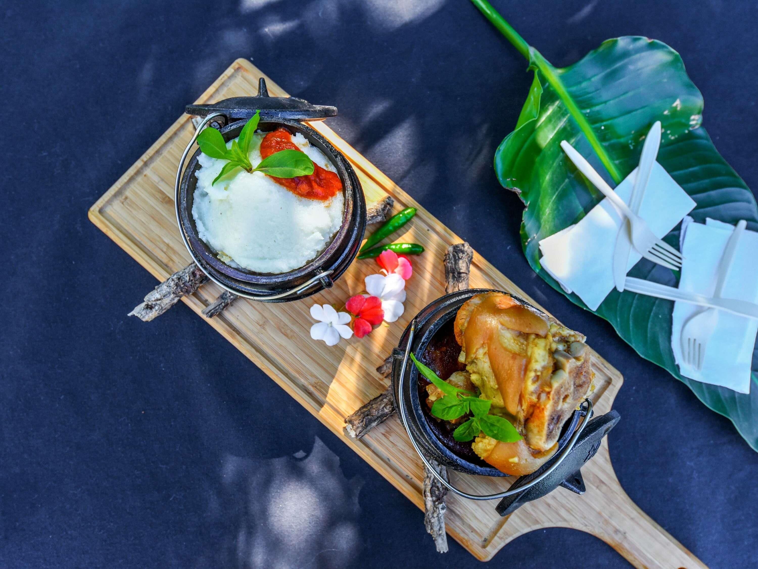 A wooden serving board with two bowls of food, a banana leaf, and a pair of white plastic utensils.