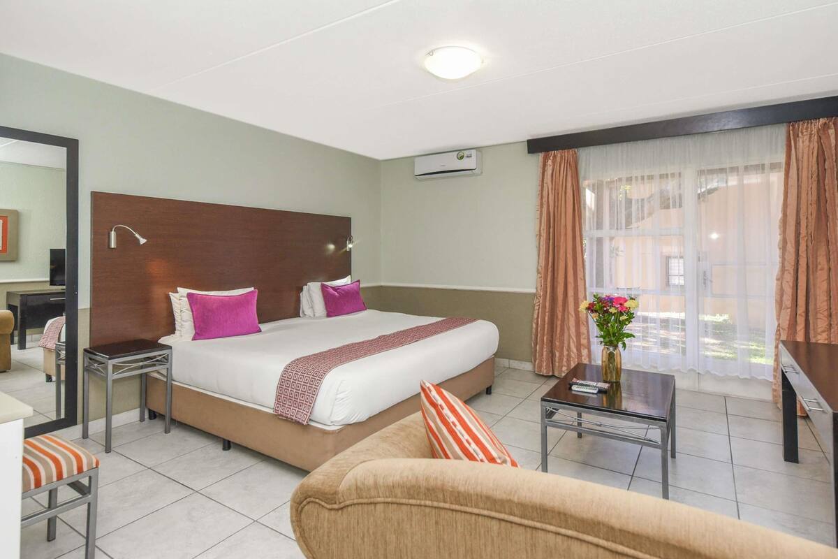 Habitación de hotel con cama, sofá, mesita, silla, escritorio, espejo y ventana con cortinas.