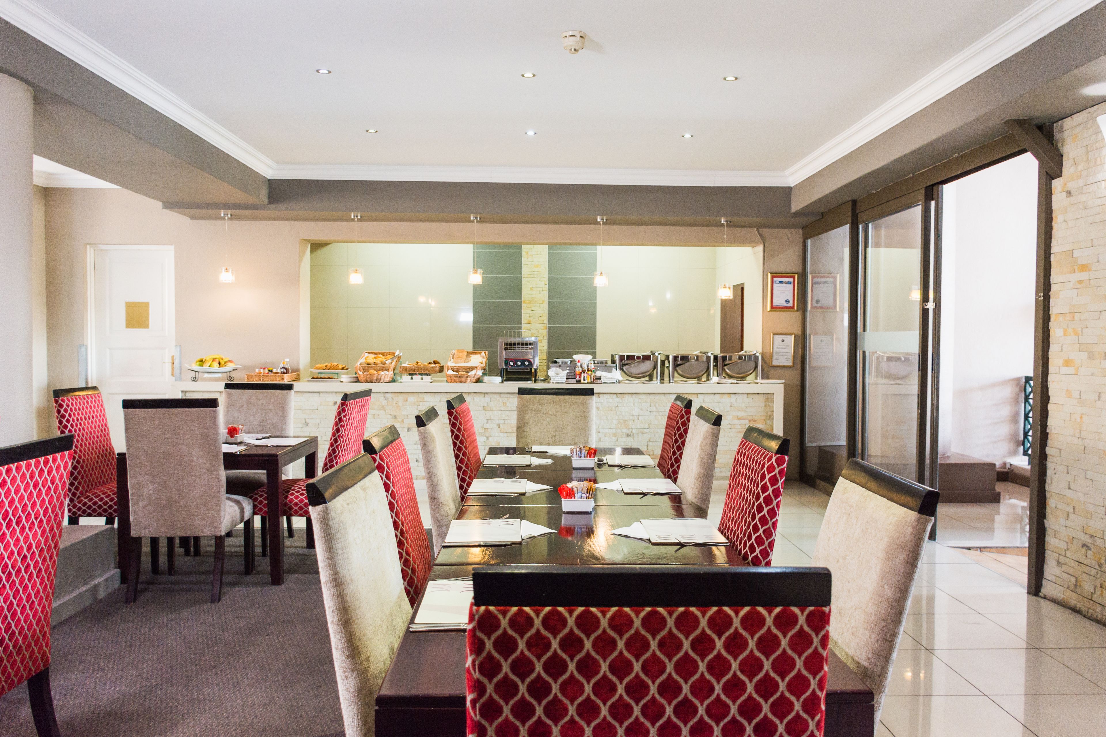 A restaurant with a long communal table, red patterned chairs, and a kitchen in the background.