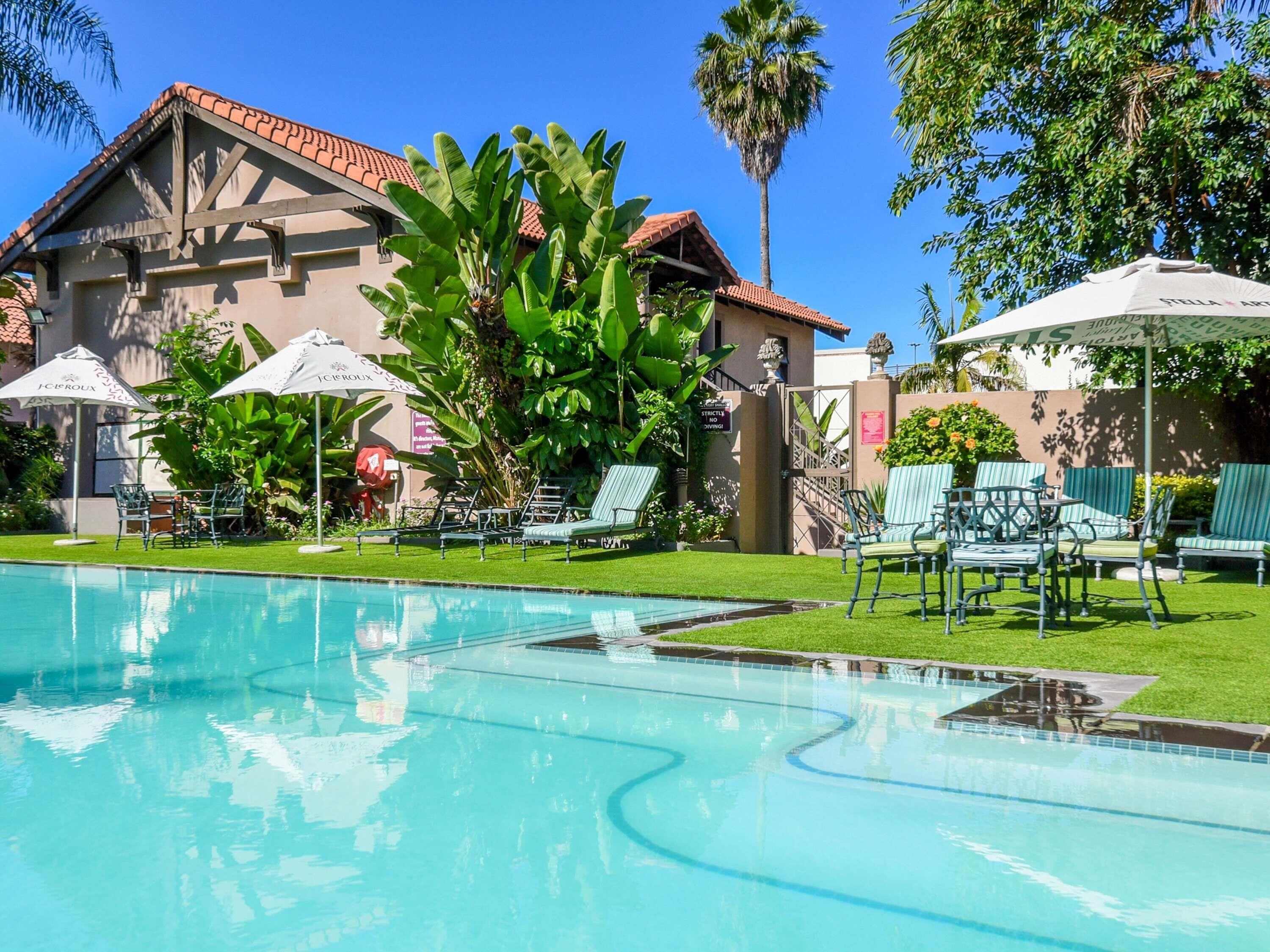 Une piscine entourée de chaises longues et de parasols, elle-même bordée d’une végétation luxuriante et d’un bâtiment doté d’un toit en tuiles.