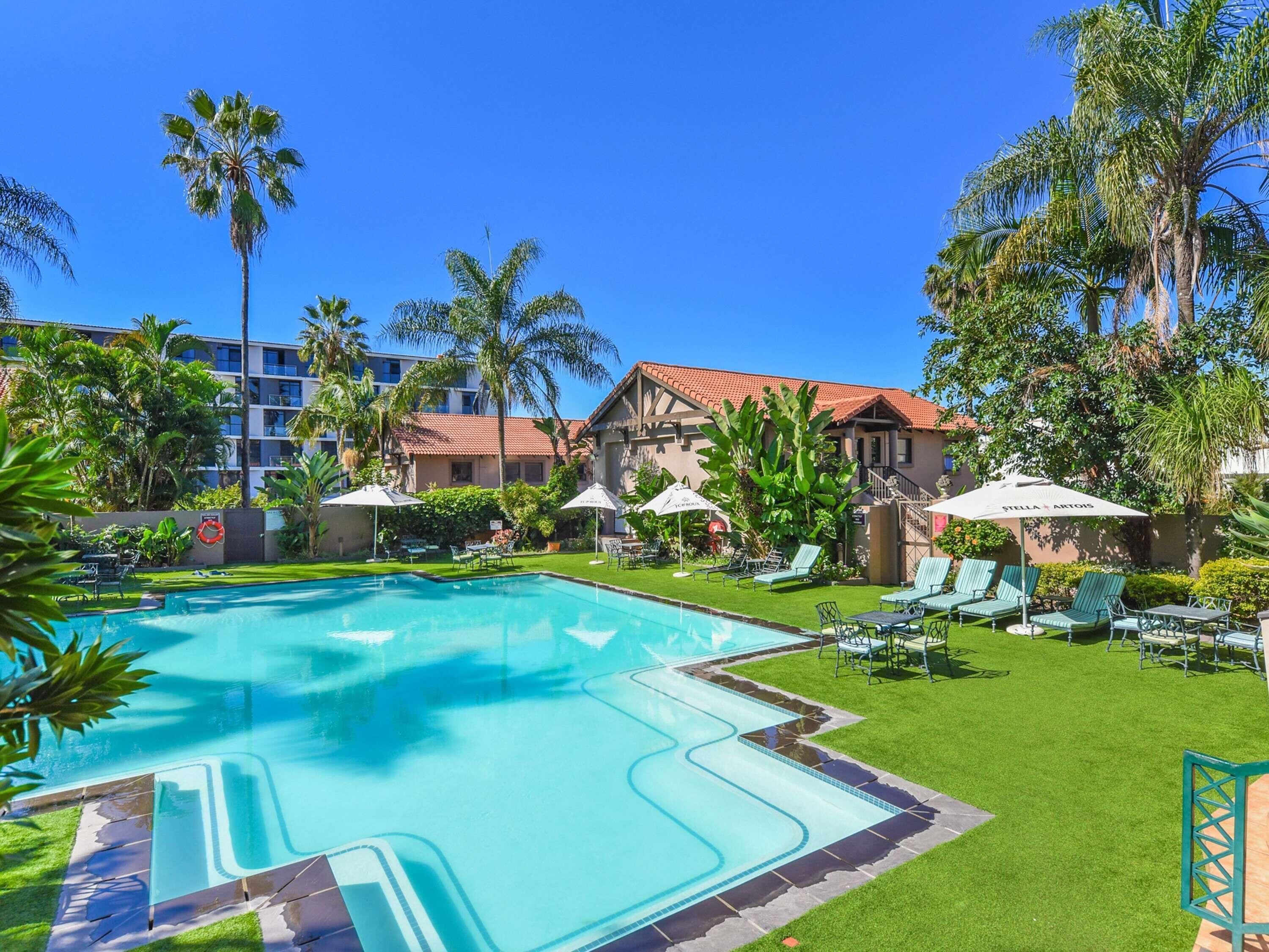 A swimming pool with lounge chairs and umbrellas, surrounded by palm trees and a building with a red roof.