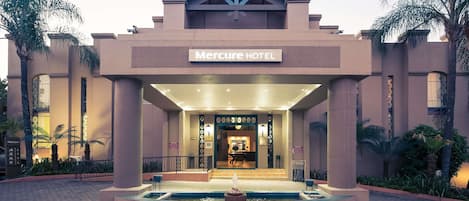 The exterior of the Mercure Hotel with a central entrance, a pool area, and palm trees.