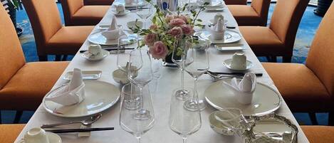 Una larga mesa de comedor preparada para una comida, con sillas a ambos lados, un florero con flores en el centro y una vista a la ciudad a través de amplios ventanales.