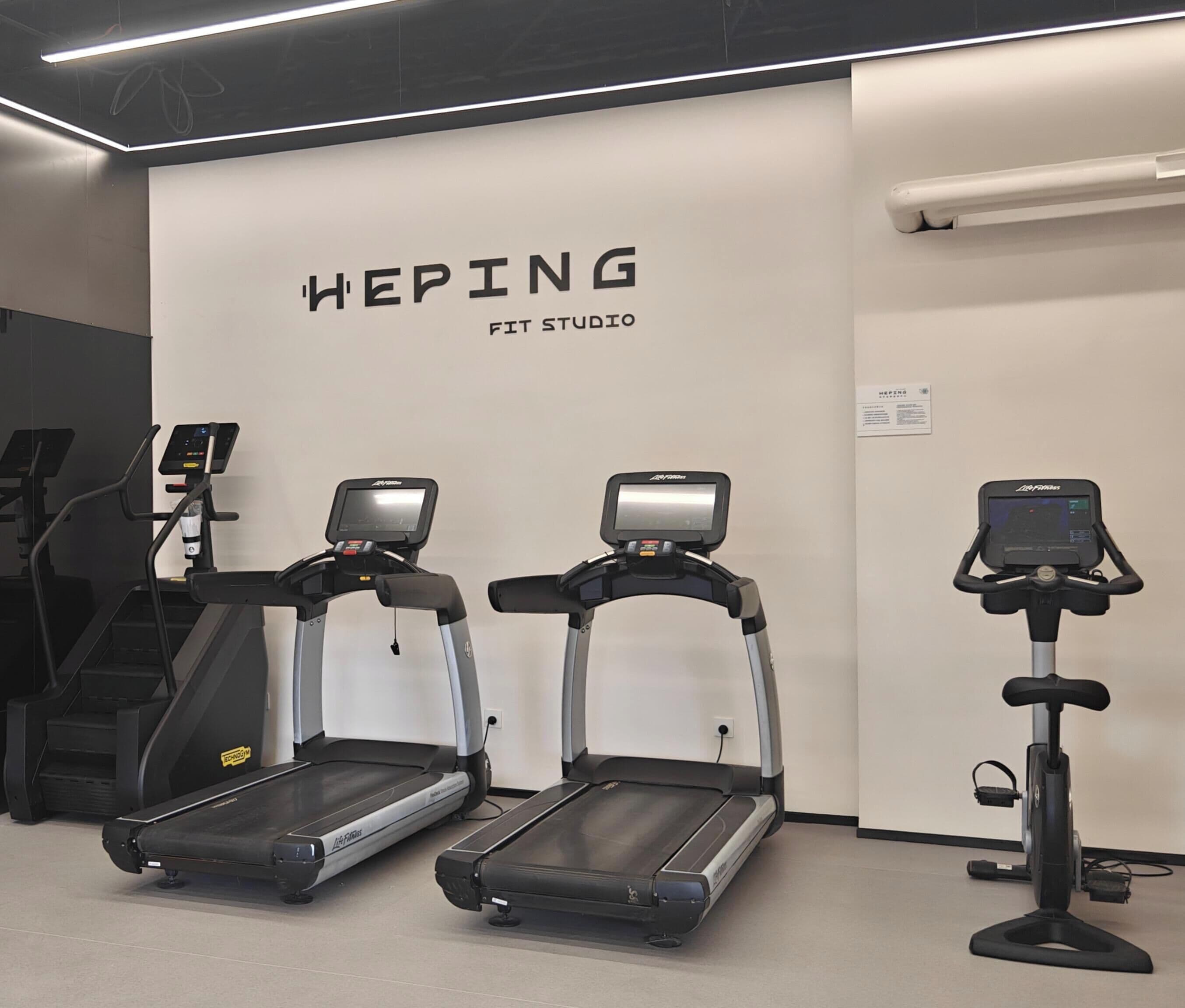 Un estudio de fitness con cintas de correr y una bicicleta estática, una pared blanca con el nombre 'HEPING FIT STUDIO' y un pequeño letrero con texto.