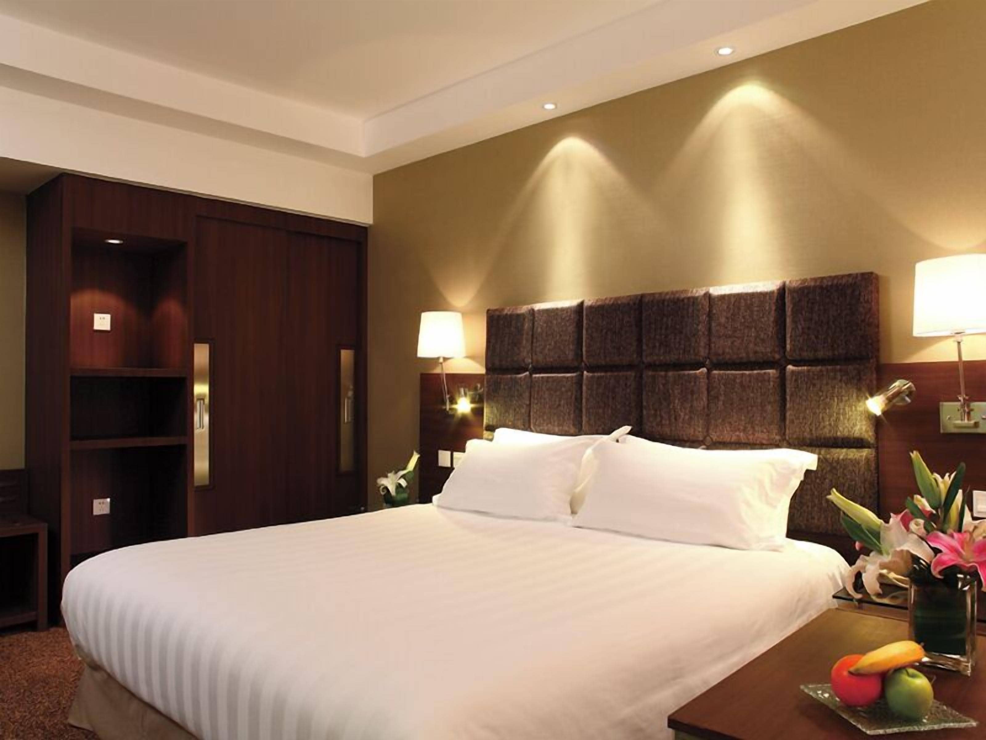 Une chambre d’hôtel avec un grand lit, une armoire, une table de chevet avec une lampe et une petite table avec des fruits.