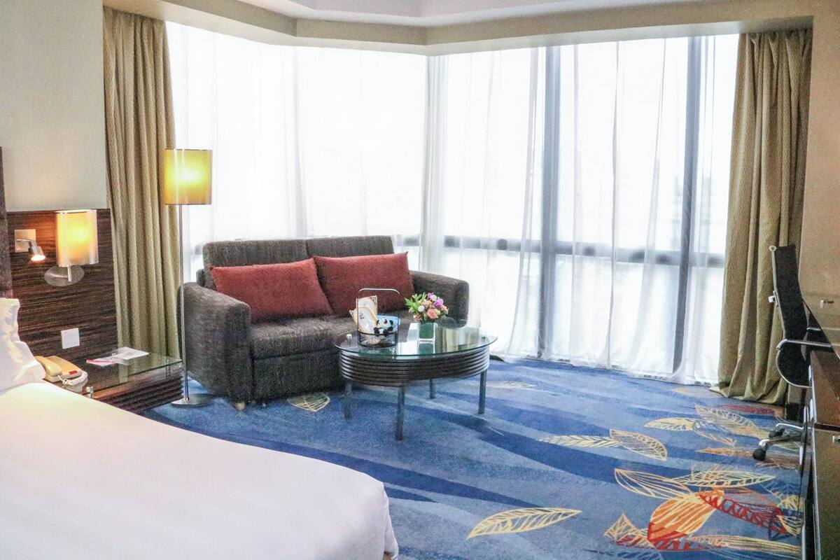 A hotel room with a large bed, a sofa with red cushions, a glass coffee table, and a view of the city through large windows.