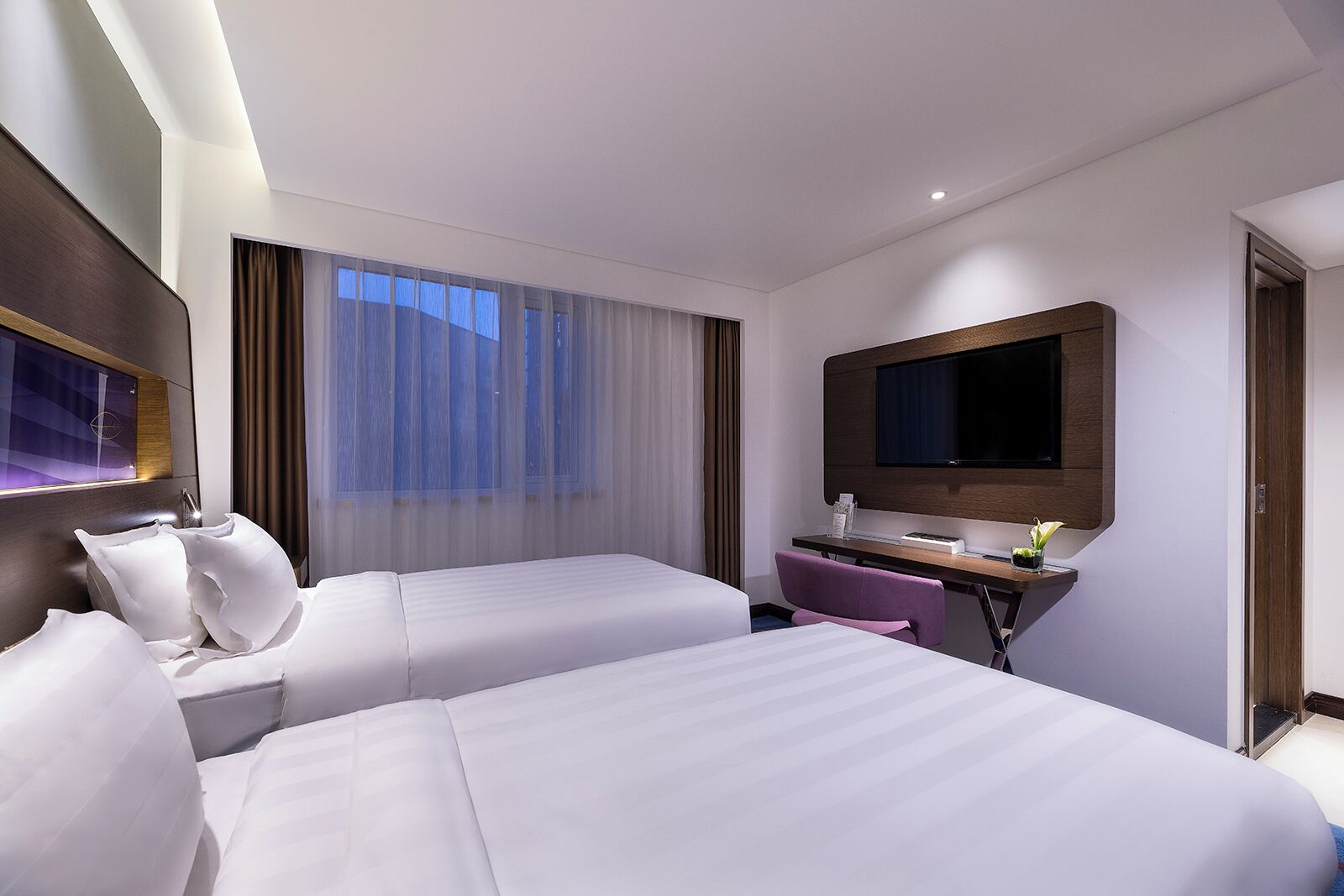 A hotel room with two beds, a desk with a computer, a TV, and a window with curtains.