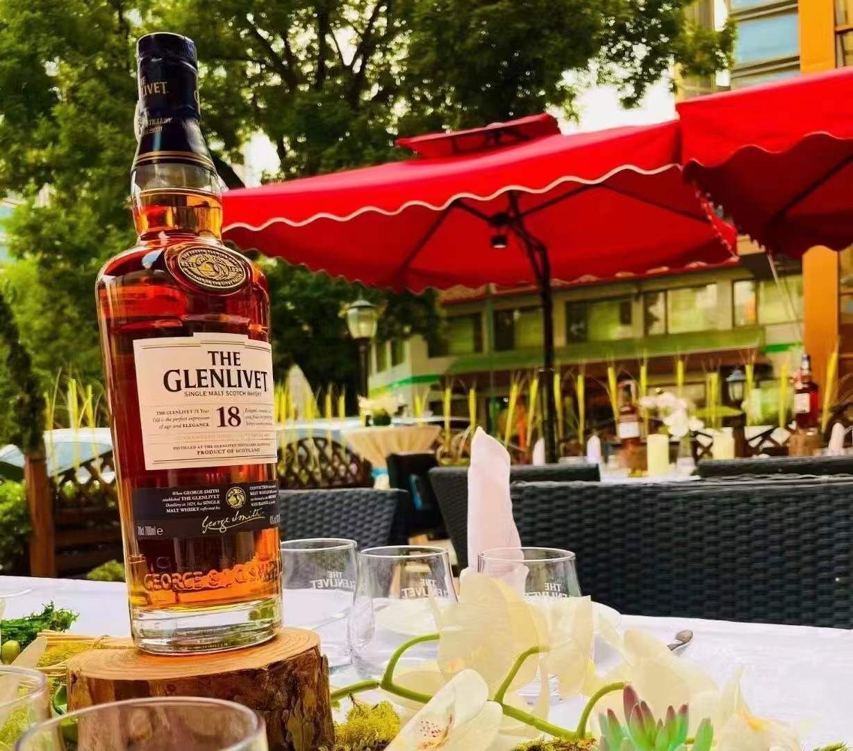 A bottle of The Glenlivet 18 single malt scotch whisky on a table with glasses and a wooden stump.