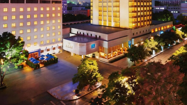 Un hotel de varios pisos con una construcción destacada en esquina, un estacionamiento y áreas verdes circundantes.
