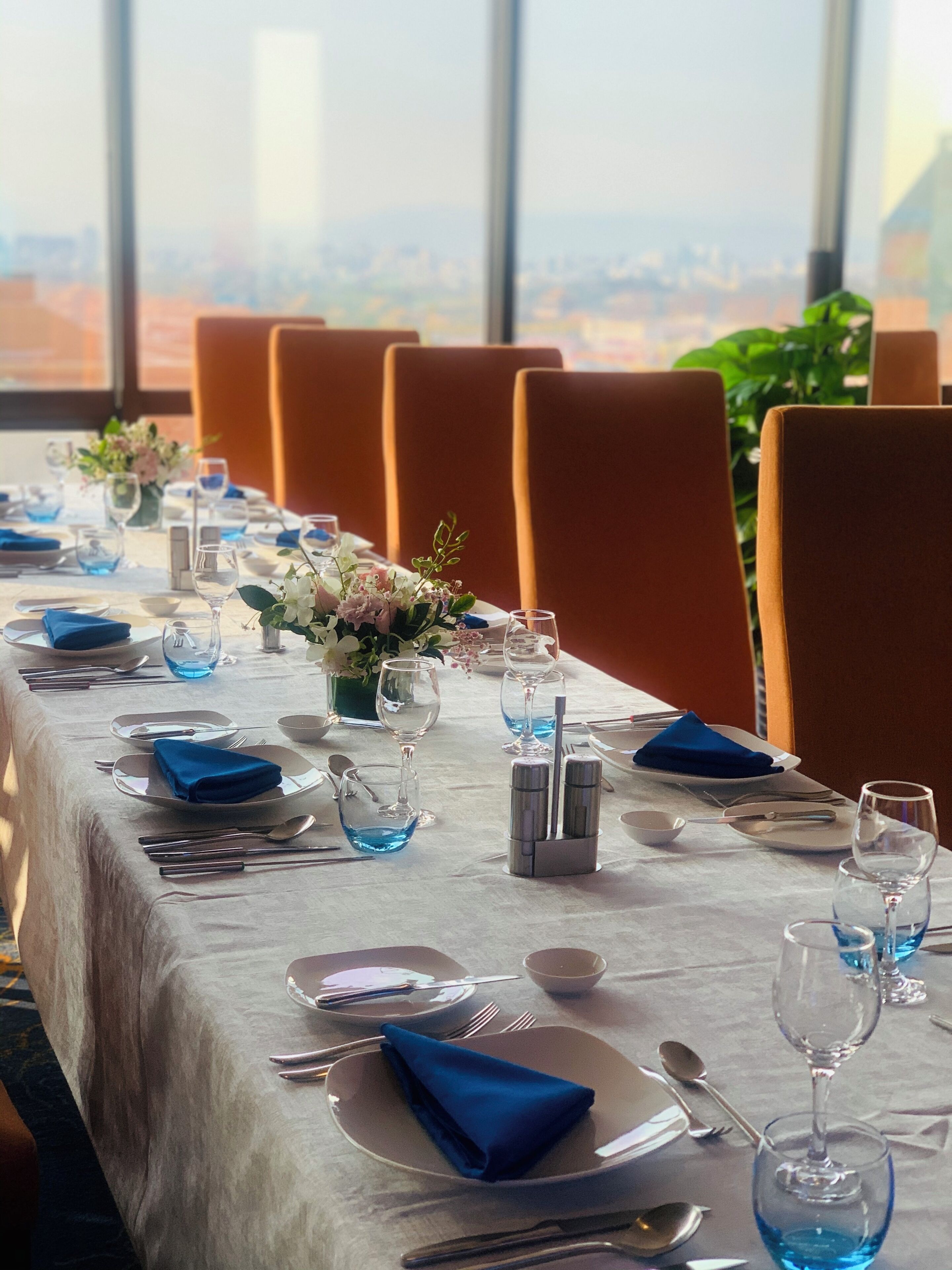 A dining table set with blue napkins, clear glasses, and silverware, with a city view through large windows.