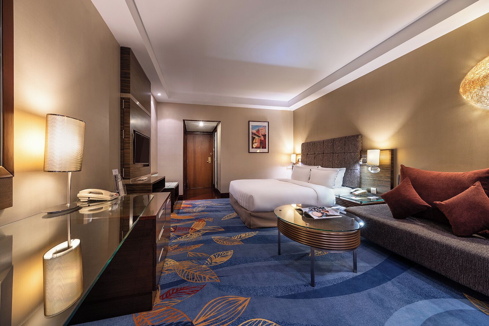 A hotel room with a bed, a sitting area with a sofa and armchair, a glass coffee table, a desk with a lamp, and a TV.