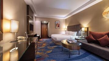 A hotel room with a bed, a sitting area with a sofa and armchair, a glass coffee table, a desk with a lamp, and a TV.