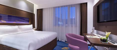 Habitación de hotel con una cama grande, un escritorio con silla, un televisor y una ventana con cortinas translúcidas.