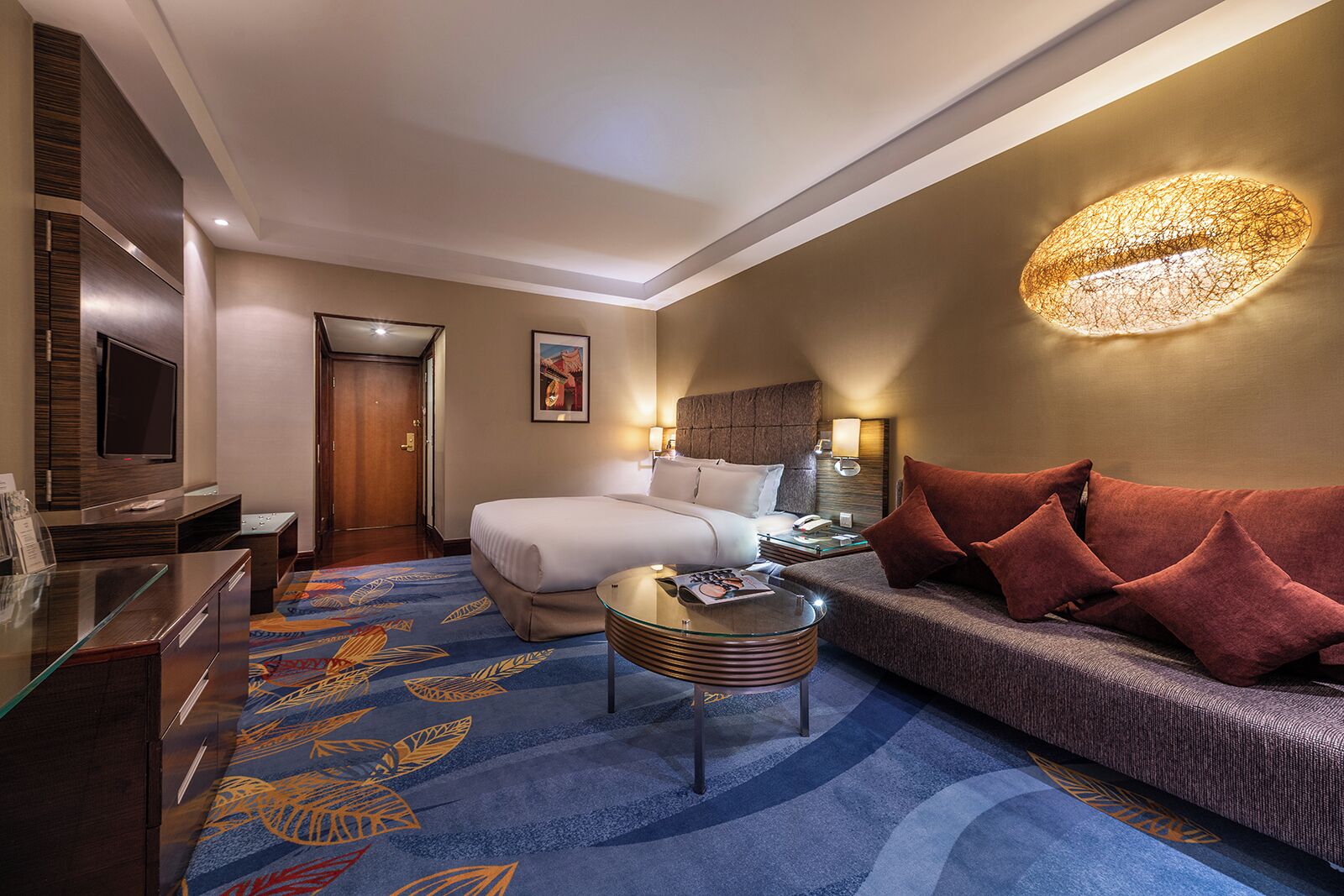 A hotel room with a bed, a sofa, a glass coffee table, a TV, and a desk with a computer.