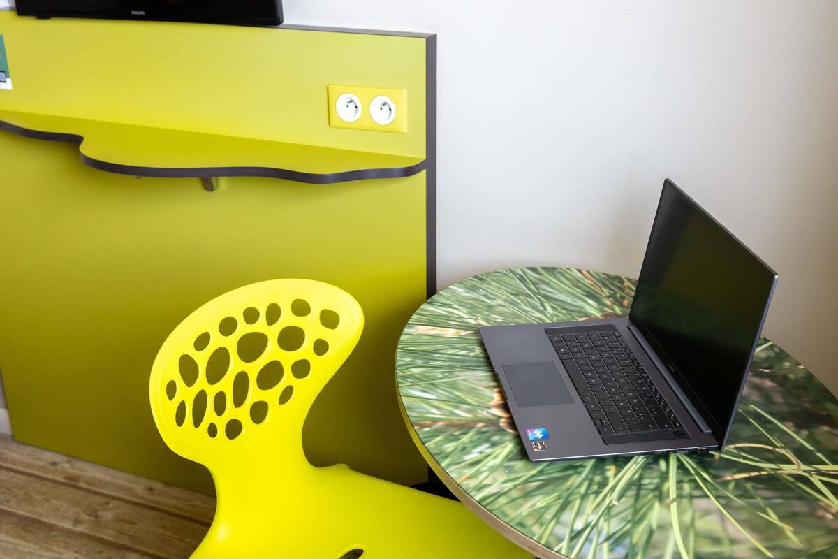 Un ordenador portátil sobre una mesa redonda con un motivo de hojas, una silla amarilla de diseño original y una pared amarilla con toma de corriente.