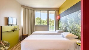 Habitación de hotel con dos camas, un televisor, una mesita y ventana con vistas a árboles.