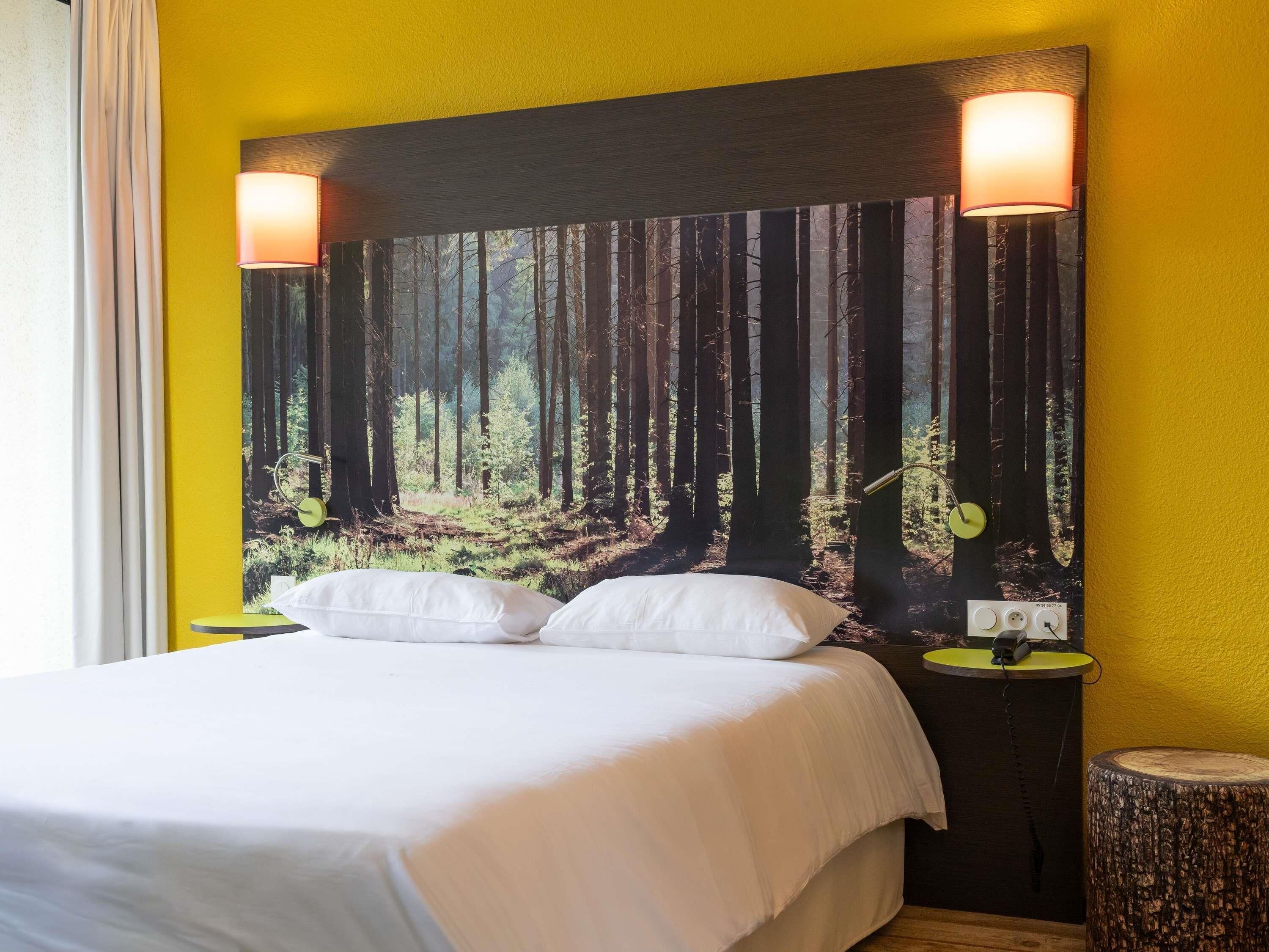 A bed with white bedding, two bedside lamps, a wall-mounted power outlet, and a large forest-themed artwork.