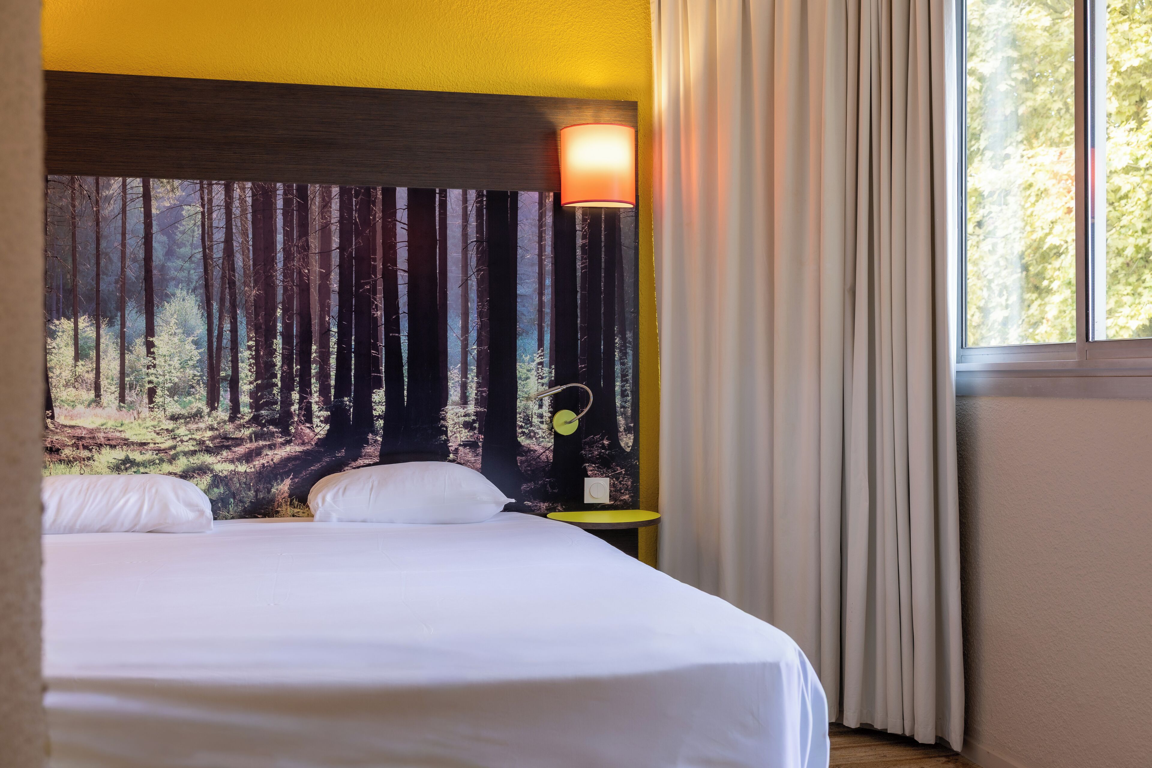 Habitación de hotel con cama, lámpara de pared, mesita de noche y ventana con cortinas.
