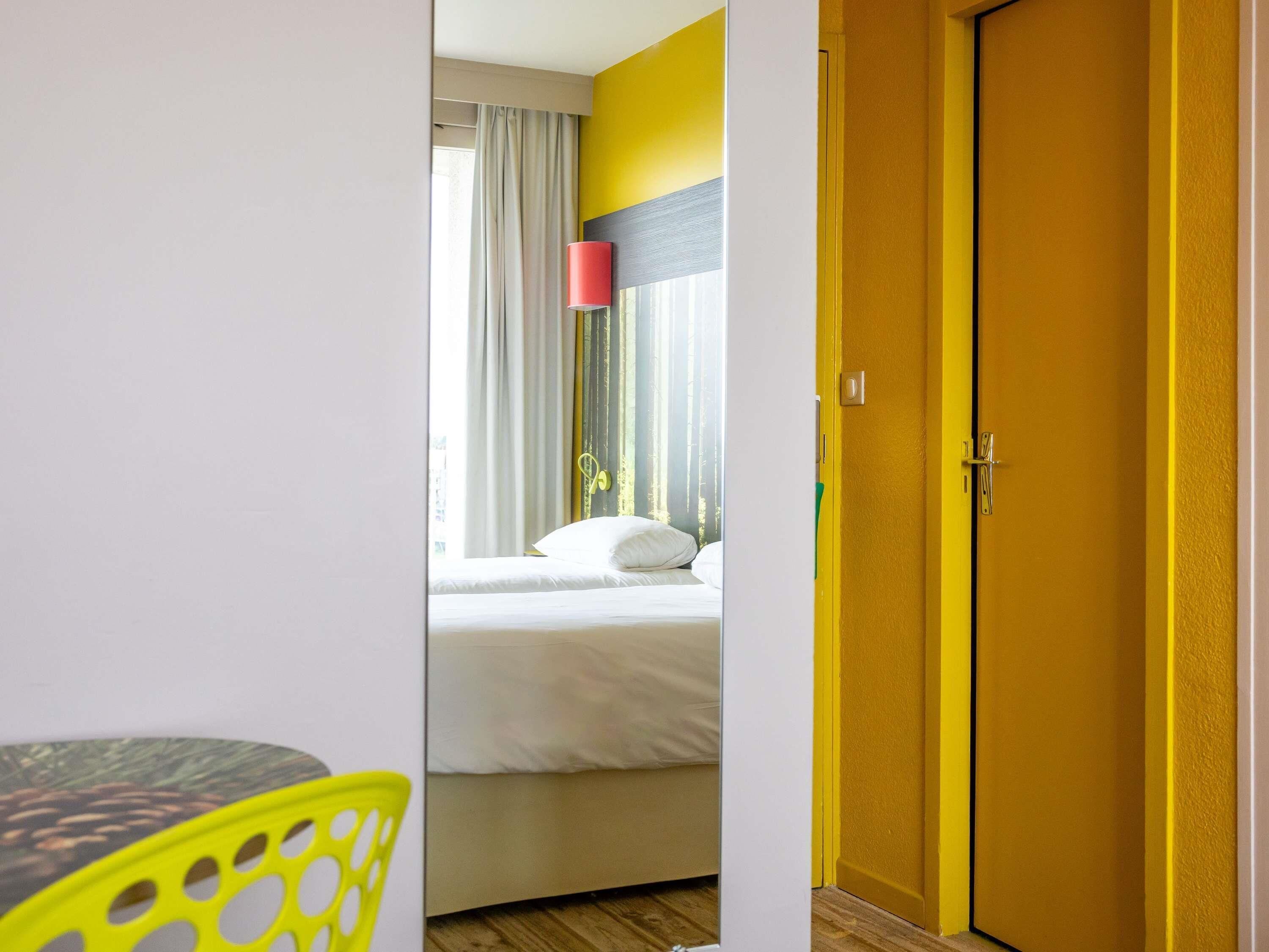 A hotel room with a bed, a yellow door, and a window with curtains.