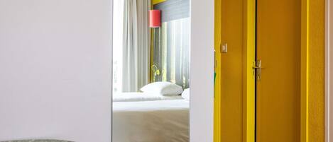 A hotel room with a bed, a yellow door, and a window with curtains.