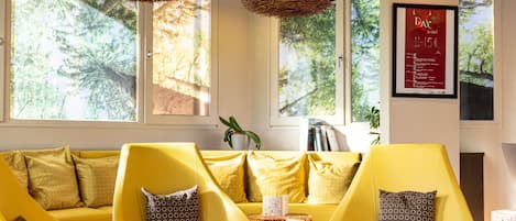 A modern living room with yellow sofas and armchairs, a round coffee table, and large windows with a view of trees.
