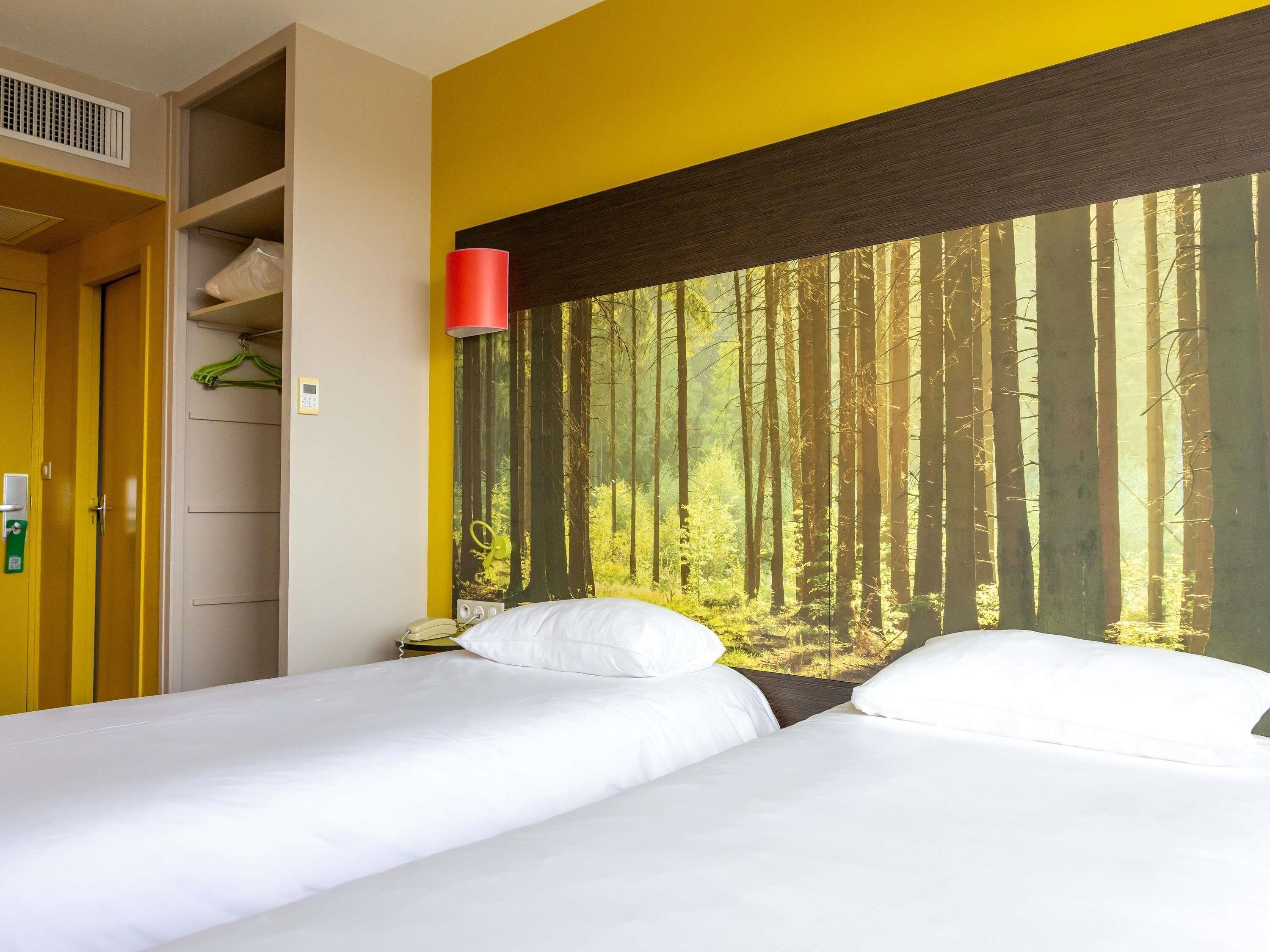 Hotellihuone, jossa on kaksi sänkyä, keltainen seinä, suuri metsäaiheinen seinämaalaus ja punainen lamppu.