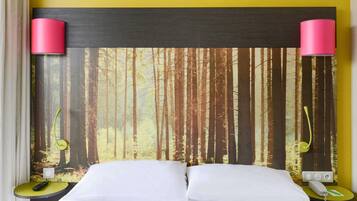 Una habitación de hotel con una cama, cabecera con papel tapiz de bosque, dos lámparas de noche y una mesita de noche con un teléfono y control remoto.