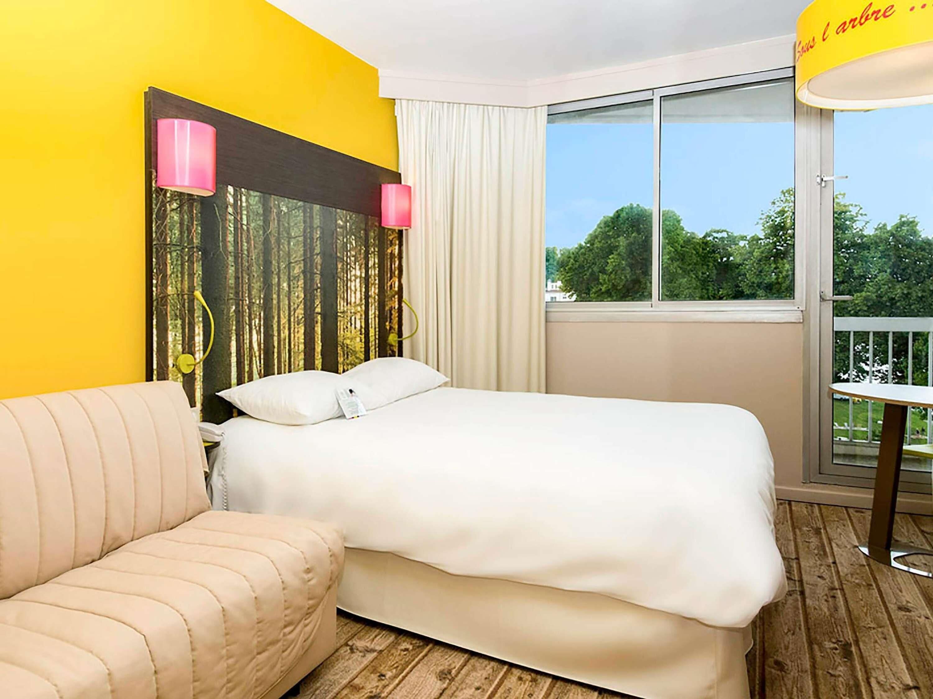 Een hotelkamer met een bed, een bank, een raam met uitzicht op bomen en een gele muur.