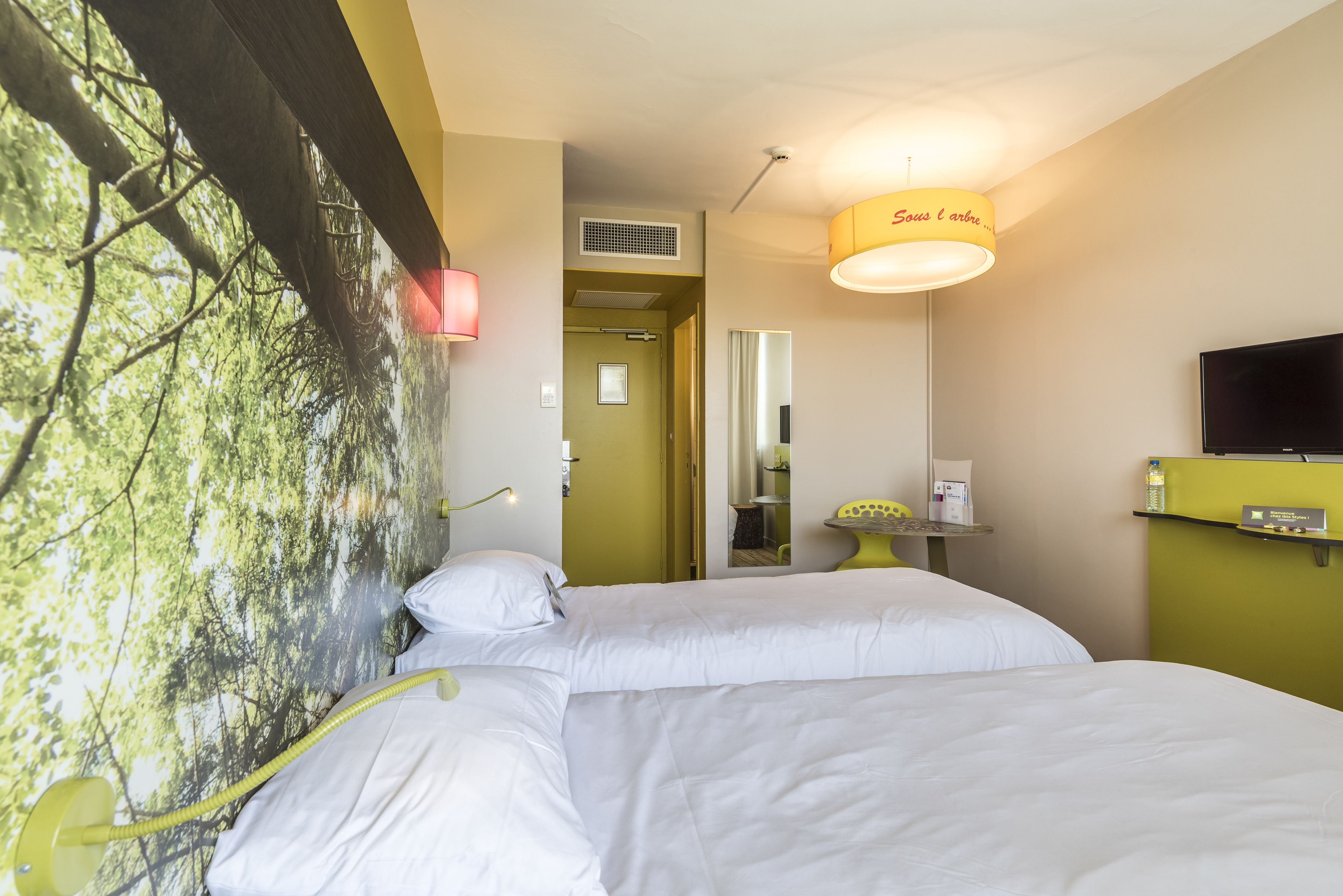 A hotel room with two beds, a television, a mirror, and a wall-mounted heater.