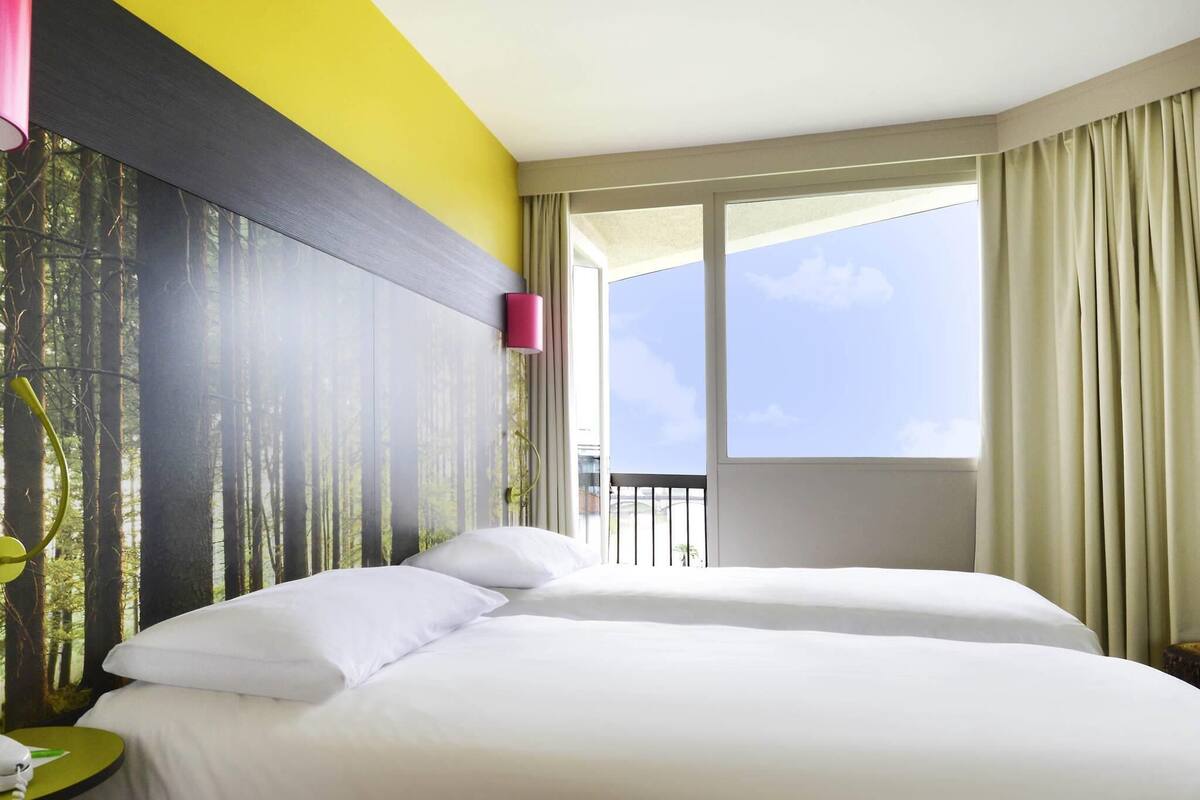 A hotel room with two beds, a large window with curtains, a nightstand with a phone, and a wall with a forest mural.