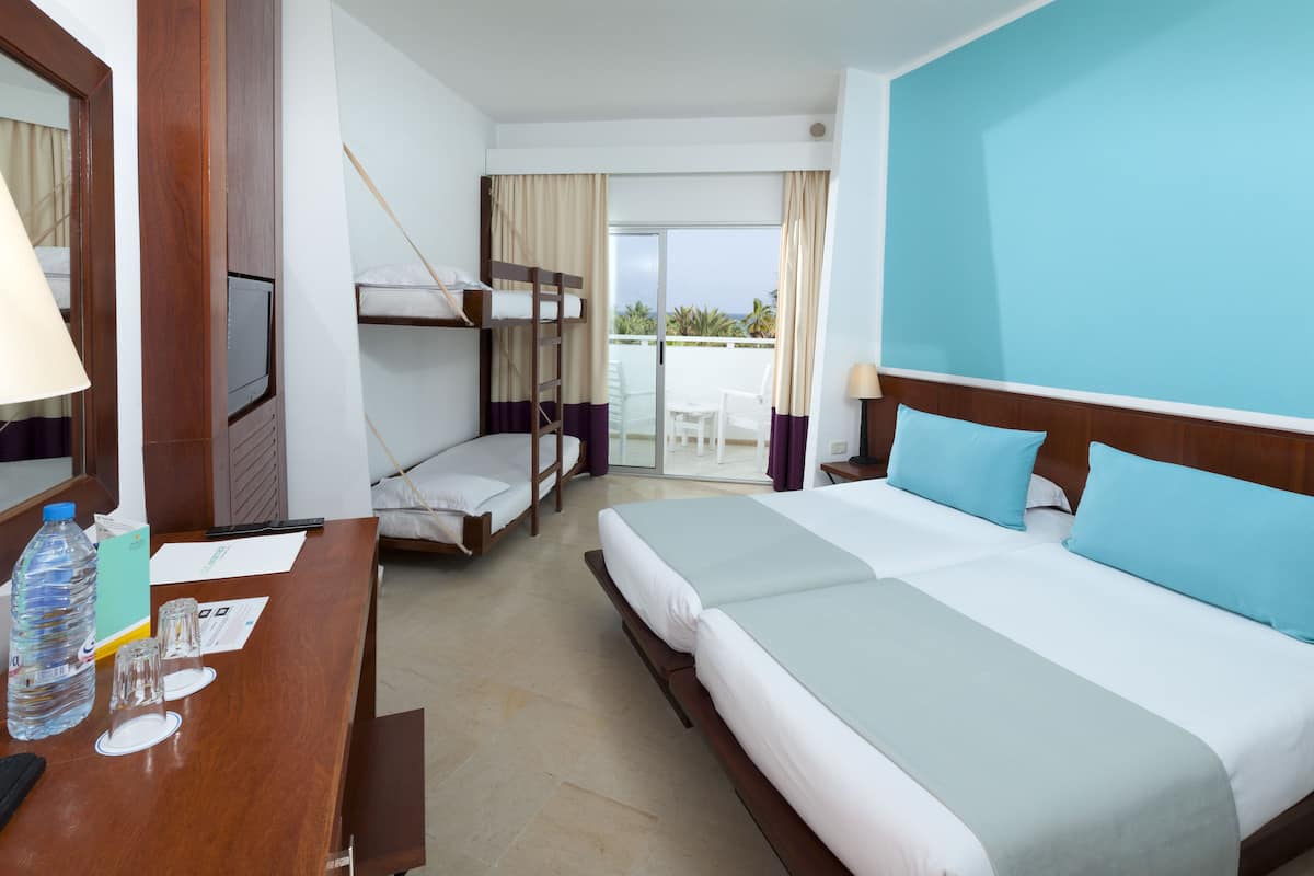 A hotel room with two beds, a desk with a lamp, a television, and a balcony with a view.